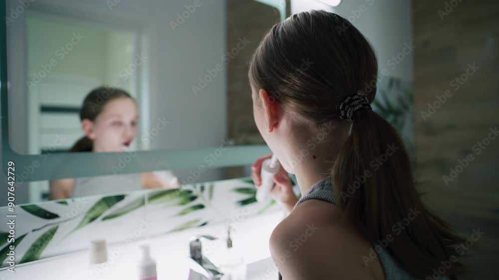 Young girl looks at mirror in bathroom, brushes her teeth. Pretty teenager stands in front of sink and uses electrical toothbrush before bedtime. Evening bath routine. Concept of lifestyle. Close up. Young girl looks at mirror in bathroom, brushes her teeth. Pretty teenager stands in front of sink and uses electrical toothbrush before bedtime. Evening bath routine. Concept of lifestyle. Close up.