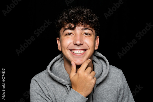 Portrait of young man about 25 years old with curly hair, holds hand on chin and smiles at camera. Isolated on black studio background photo