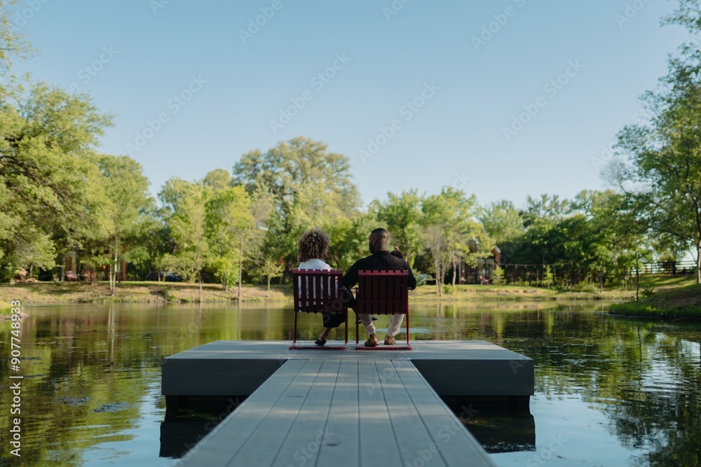 Obraz premium Back shot of couple sitting on chairs at the end of a dock overlooking a serene lake on a sunny day