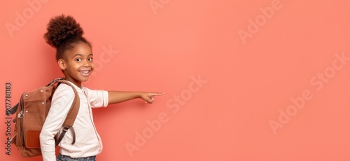 Smiling African American girl with puffy hair, pointing to the side, wearing a white shirt and brown backpack against a pink background.