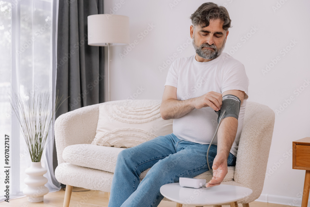 Handsome grey-haired senior man in casual outfit sitting on couch at home, checking blood pressure on his own. Pensioner using modern tonometer, experiencing hypertension, copy space Handsome grey-haired senior man in casual outfit sitting on couch at home, checking blood pressure on his own. Pensioner using modern tonometer, experiencing hypertension, copy space