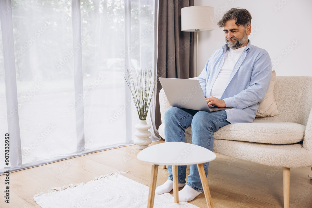Smiling mature man using laptop sitting on armchair in living room. Happy confident senior male adult resting at home working on pc, typing on keyboard, full body length, copy space Smiling mature man using laptop sitting on armchair in living room. Happy confident senior male adult resting at home working on pc, typing on keyboard, full body length, copy space