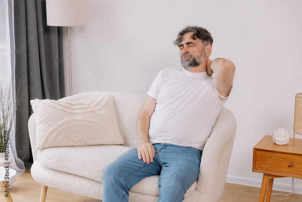 A senior man is sitting indoors, visibly suffering from neck pain. He is holding his neck with a pained expression, indicating discomfort, copy space for your advertising A senior man is sitting indoors, visibly suffering from neck pain. He is holding his neck with a pained expression, indicating discomfort, copy space for your advertising