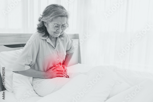 Fotografie Elderly Asian woman had stomach pain with pained expression on her face while sleeping on the bed, holding her stomach with her hands in excruciating pain for no apparent reason