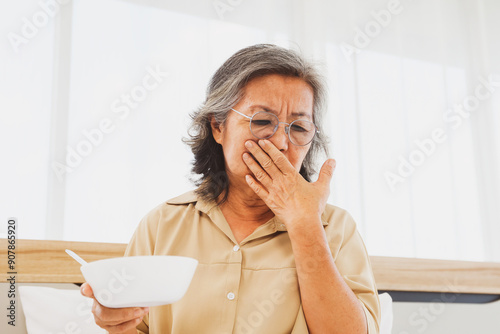 Sick elderly asian woman holds breakfast bowl with an expression that she cannot eat use your hand to cover your mouth tired eating boring unpalatable bland food lack flavor and monotonous.