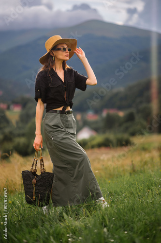 Tableau sur toile Fashionable confident woman wearing trendy summer hat, sunglasses, linen shirt, green maxi skirt, silver cowboy boots, holding black wicker straw bag, walking outdoor