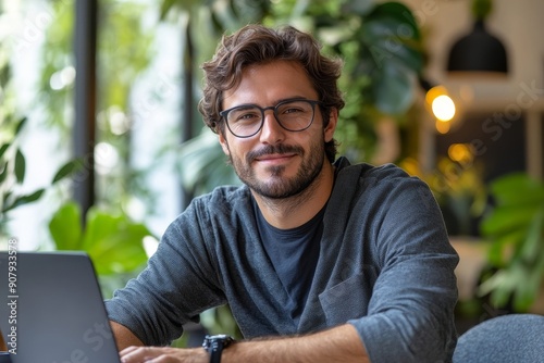 Portrait of a young man in glasses sitting at a desk in the office and working on a laptop, looking and smiling at the camera, Generative AI