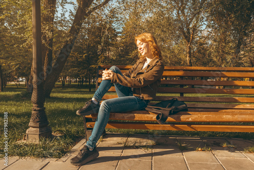 Smiling Redhead Woman in Brown Jacket Enjoying Autumn Day in Sunlit Park with Golden Foliage