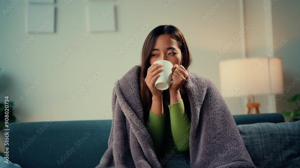Young woman wearing a knitted sweater sitting at home on the sofa wrapped in a plaid drinking hot coffee or tea Young woman wearing a knitted sweater sitting at home on the sofa wrapped in a plaid drinking hot coffee or tea
