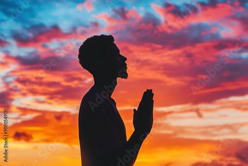 Prayer concept. Silhouette of a young man in a praying pose. Set against a vibrant sunset sunrise sky. Clasped hands. Also related to gospel, testimony, deliverance, providence, mercy, ai © ImronDesign