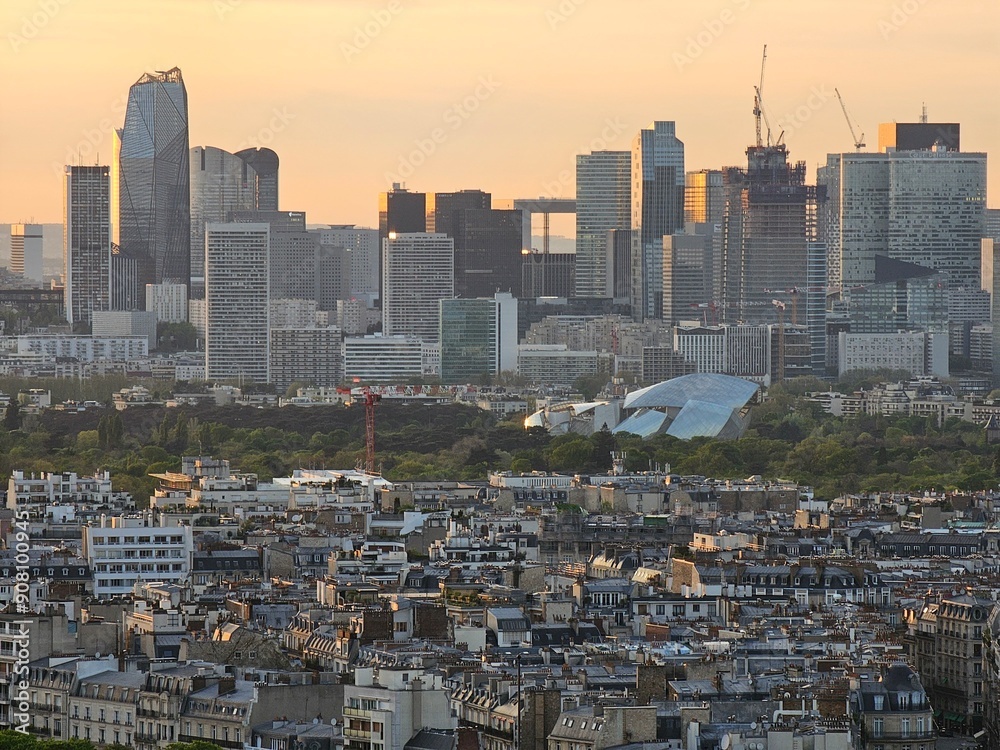 Fototapeta premium Paris, France - April 12, 2024: Aerial view to the city of Paris, during the golden hour between modern skyscrapers and old buildings. Beautiful sunset over the city reflected on the buildings.
