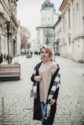 A woman wearing a checkered jacket and scarf stands on a cobblestone street. She is smiling and she is enjoying the day