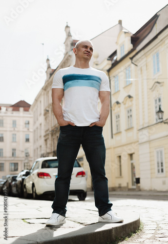 A man stands on a sidewalk in front of a parking meter. He is wearing a white shirt and blue jeans