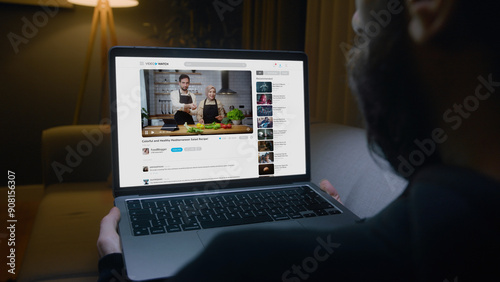 Man lying on sofa watching a food blogger couple's video on a laptop, learning how to prepare a healthy Mediterranean salad in living room at night.	
 photo