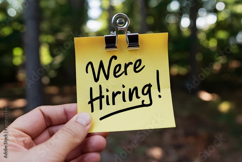 Hand holding yellow sticky note with Were Hiring against blurred trees photo