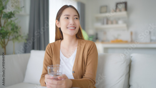 Free Download Happy young Asian woman drinking water while sitting on the sofa in the living room - GFXLoad