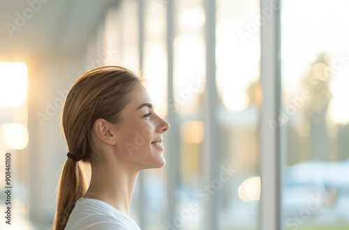 A profile view of a smiling woman looking out of large windows with a serene and hopeful expression in a bright modern setting. Free Download A profile view of a smiling woman looking out of large windows with a serene and hopeful expression in a bright modern setting. - GFXLoad