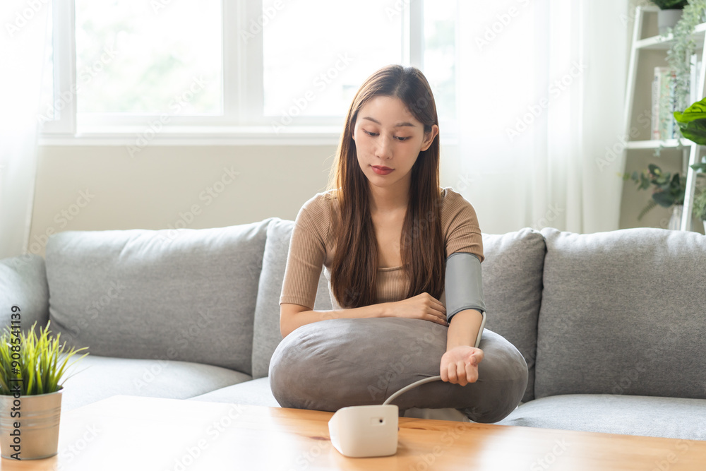 Hypotension, asian young woman sitting on couch checking blood high pressure and heart rate with digital pressure monitor machine making self check up on her arm at home. Healthcare and medical. Hypotension, asian young woman sitting on couch checking blood high pressure and heart rate with digital pressure monitor machine making self check up on her arm at home. Healthcare and medical.