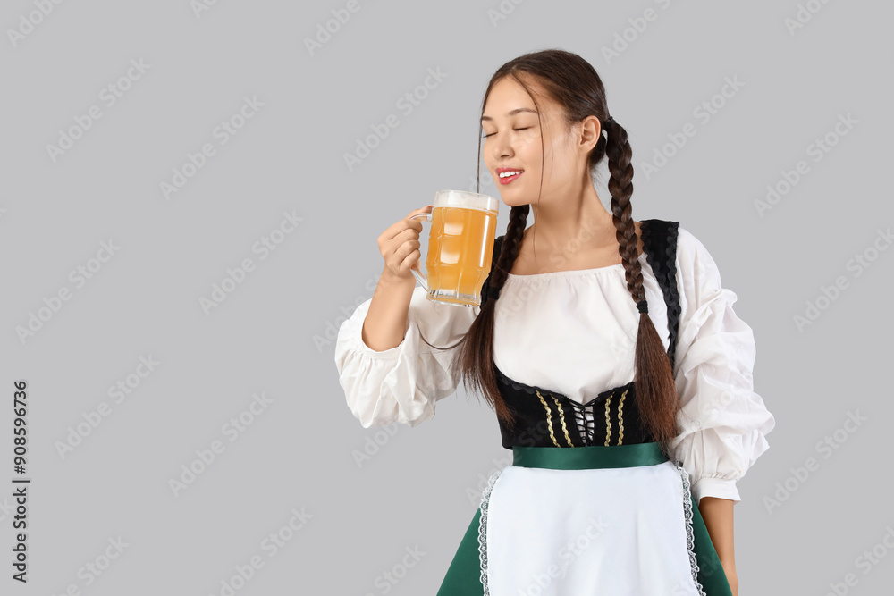 Beautiful young happy Asian Octoberfest waitress with mug of beer on grey background Beautiful young happy Asian Octoberfest waitress with mug of beer on grey background