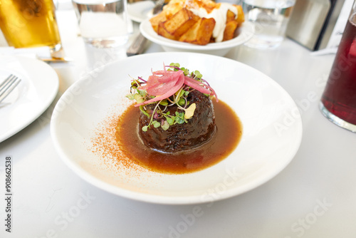 Braised beef cheek with sauce, garnished with herbs on a white plate next to a beer and a wine cocktail and tapas patatas bravas on a table in a restaurant