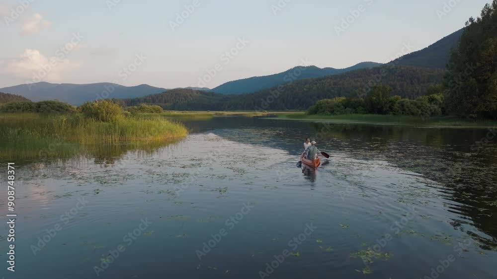 Active couple taking a canoe ride across the beautiful mountain lake, enjoying summer nature on a lakeshore vacation, aerial rear view shot. Active couple taking a canoe ride across the beautiful mountain lake, enjoying summer nature on a lakeshore vacation, aerial rear view shot.