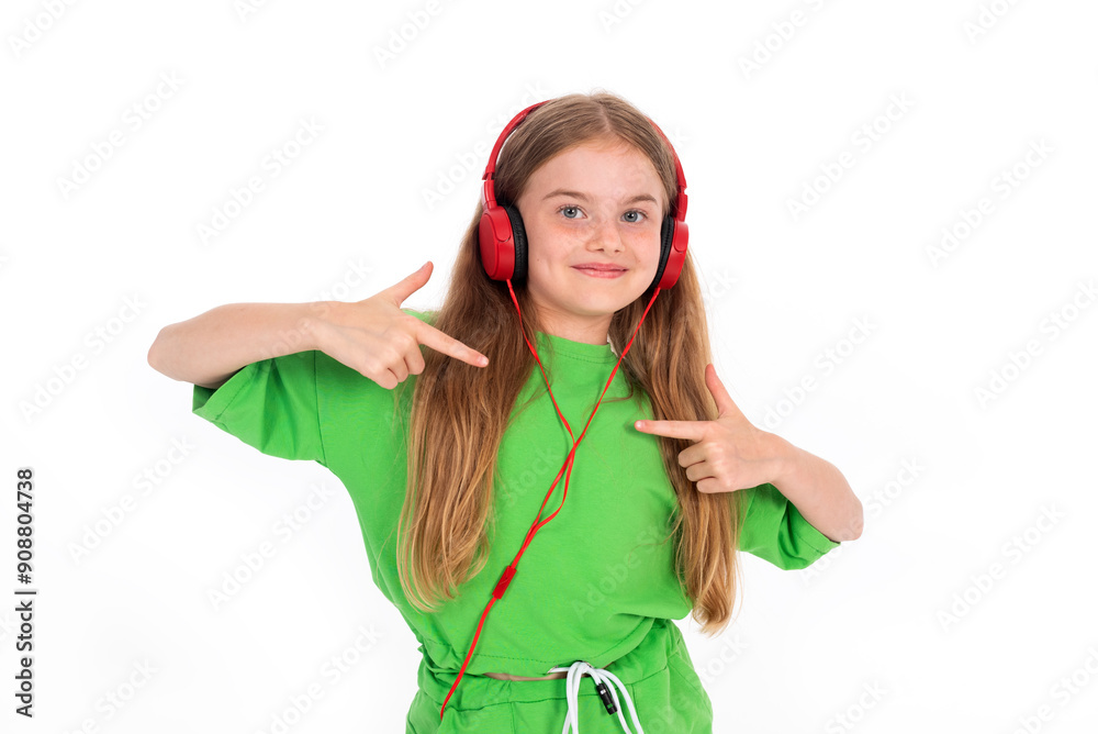 Smiling young girl in a green outfit wearing red headphones, pointing at them, standing against a white background. Smiling young girl in a green outfit wearing red headphones, pointing at them, standing against a white background.