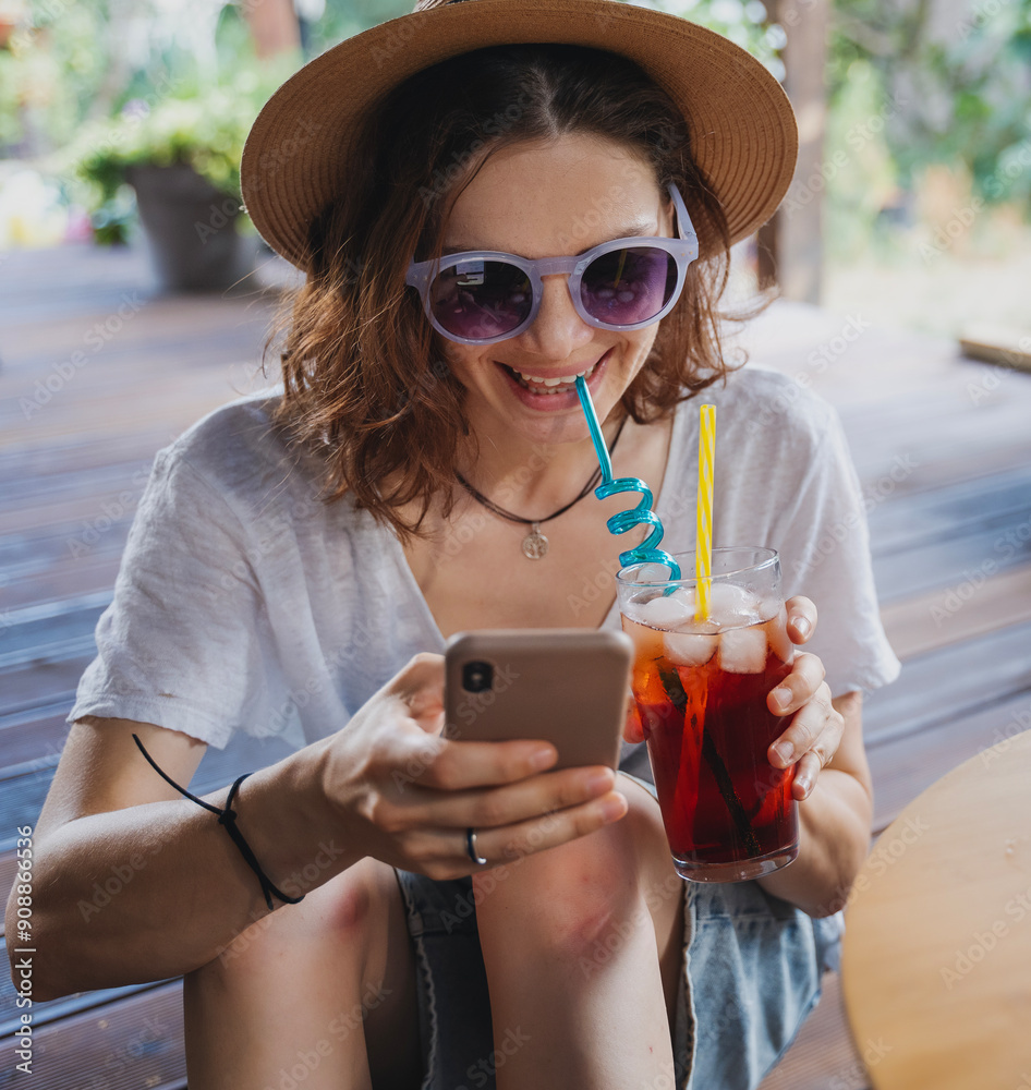 Young cheerful woman in hat with refreshing cocktail with ice and smartphone in hands on summer terrace of cottage
