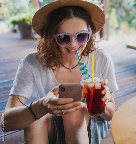 Obraz na płótnie Young cheerful woman in hat with refreshing cocktail with ice and smartphone in hands on summer terrace of cottage w salonie