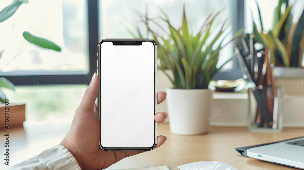 A hand holds a smartphone with a blank white screen in a modern, plant-filled office setting, suggesting technology and productivity. A hand holds a smartphone with a blank white screen in a modern, plant-filled office setting, suggesting technology and productivity.