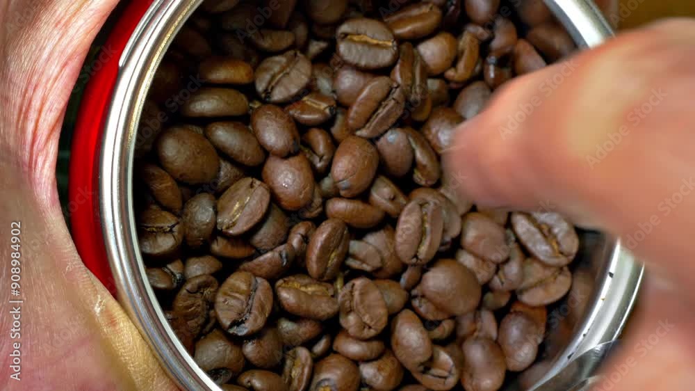 Closeup overhead shot of a hand holding a fresh tin of dark brown roasted coffee beans, while the lid is pulled off, then a finger stirs the beans. Closeup overhead shot of a hand holding a fresh tin of dark brown roasted coffee beans, while the lid is pulled off, then a finger stirs the beans.