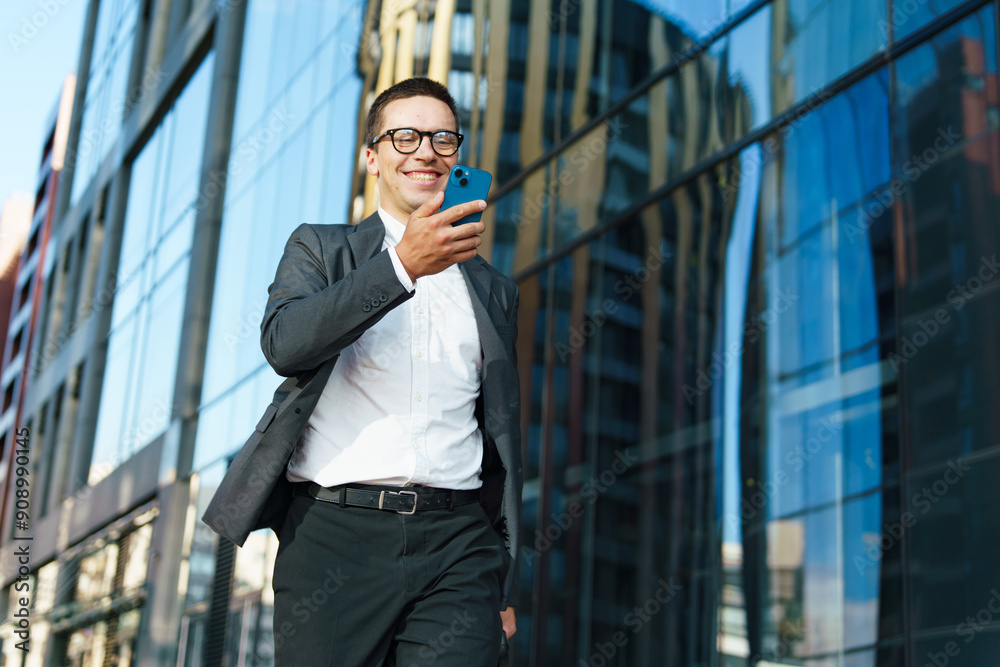 A businessman in a suit smiles while looking at his smartphone, walking in a modern urban area.