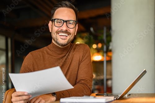 Happy male project manager with document and laptop at desk looking at camera while working in office