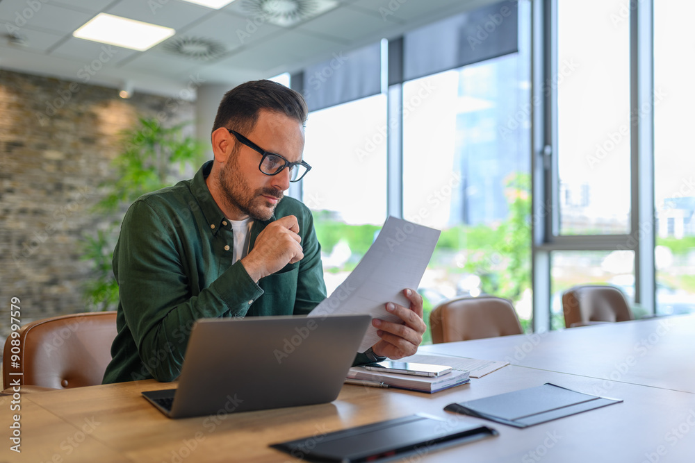 Focused young businessman in glasses reading document while working over computer at desk in office Focused young businessman in glasses reading document while working over computer at desk in office