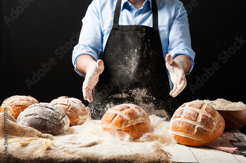 Male baker wearing black apron holding a freshly baked rustic bread splashing flour in front of a white wooden table filled with different kinds of loaves and jutte fabric