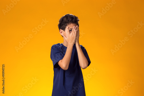 Upset kid boy covering face with palms and looking through fingers over yellow background. Emotions and bulling concept