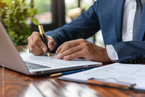 Businessman in a suit working on a laptop, writing notes on paper at a desk in a modern office, highlighting productivity and focus.