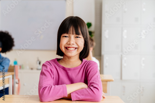 Free Download Smiling Asian schoolgirl in a classroom. Young female student with black hair sitting at a desk in a pink shirt, radiating joy and friendliness. Elementary student enjoying school life and learning - GFXLoad