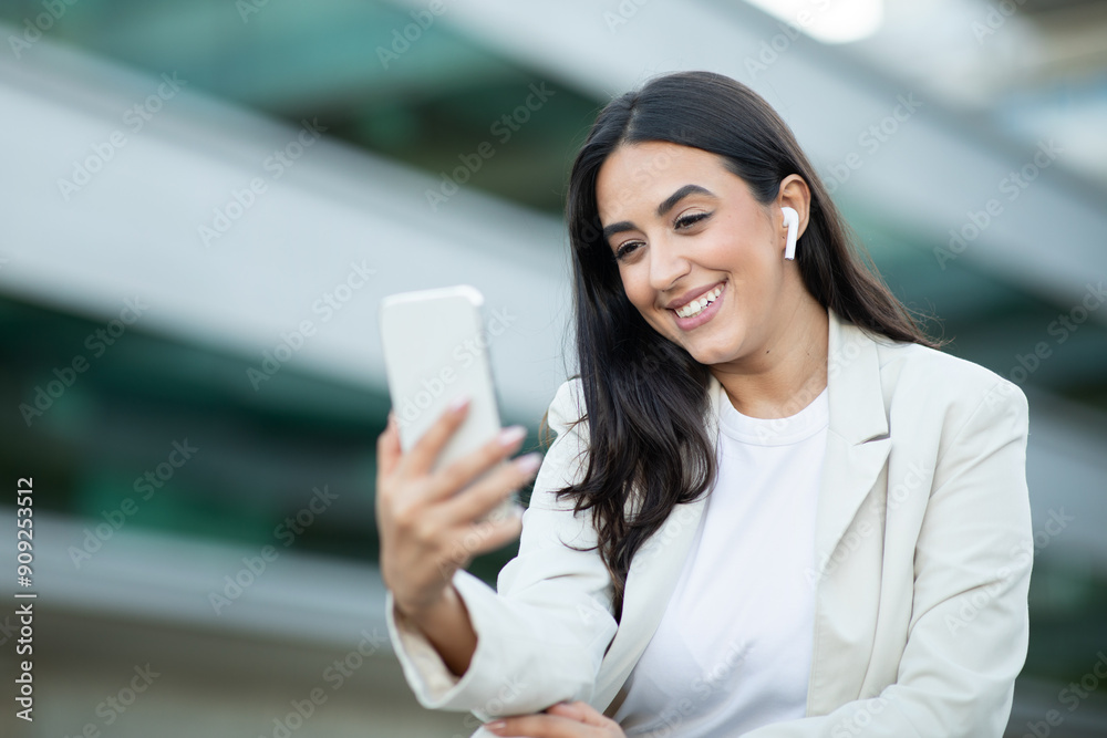 A woman with long dark hair and a white blazer smiles while she uses her smartphone. She is wearing white earbuds and is in a city setting with modern buildings in the background. A woman with long dark hair and a white blazer smiles while she uses her smartphone. She is wearing white earbuds and is in a city setting with modern buildings in the background.