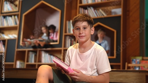 Caucasian boy looking at camera while group of smart students sitting at library. Child studying, learning, reading from novel or textbook while children talking, chitchat about education. Erudition. photo