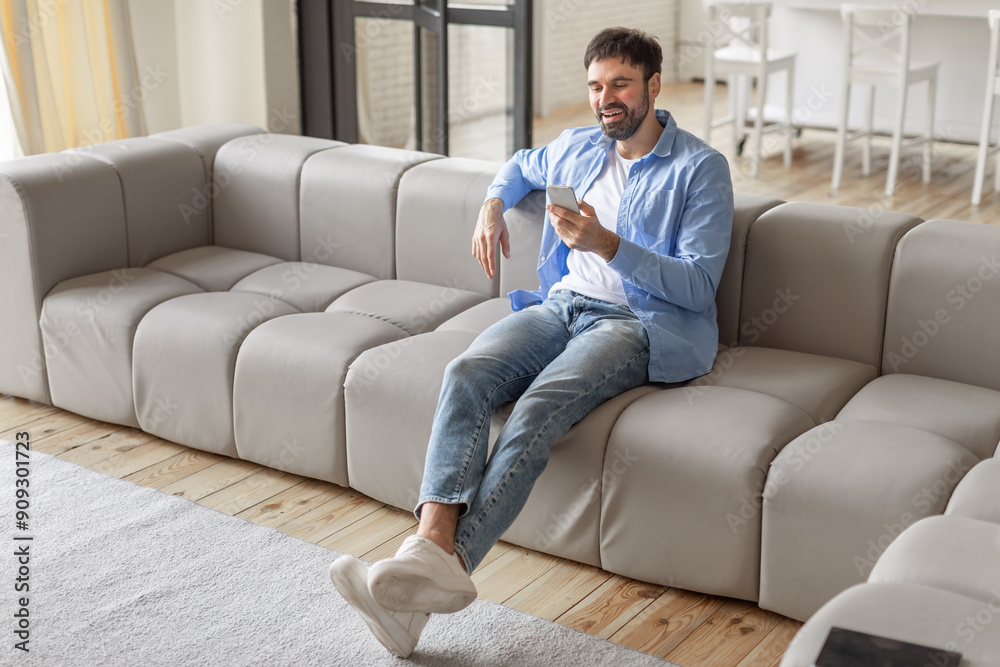 A man, dressed in jeans and a light blue shirt, sits on a light grey sofa in a modern living room. He is smiling as he uses his smartphone, copy space A man, dressed in jeans and a light blue shirt, sits on a light grey sofa in a modern living room. He is smiling as he uses his smartphone, copy space