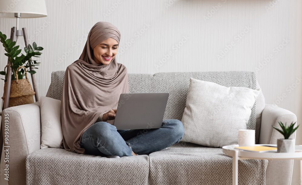 Modern arab girl in hijab using laptop at home, working remotely while sitting on couch in living room Modern arab girl in hijab using laptop at home, working remotely while sitting on couch in living room