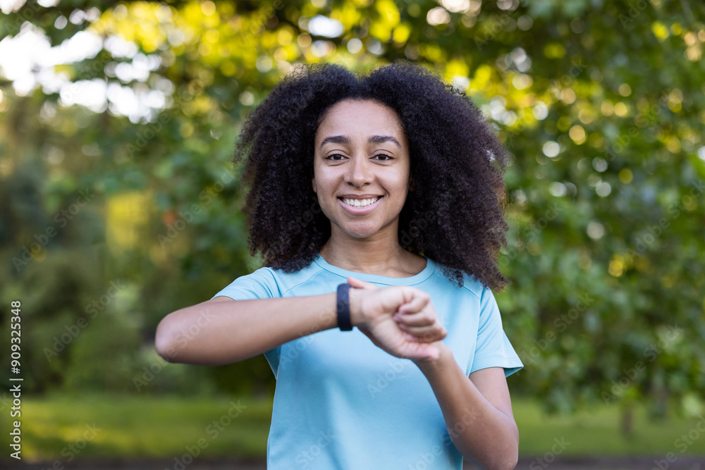 Young African American athlete checking smartwatch in green outdoor park. Smiling woman wearing sportswear and monitoring fitness progress. Concept of healthy lifestyle, technology Young African American athlete checking smartwatch in green outdoor park. Smiling woman wearing sportswear and monitoring fitness progress. Concept of healthy lifestyle, technology