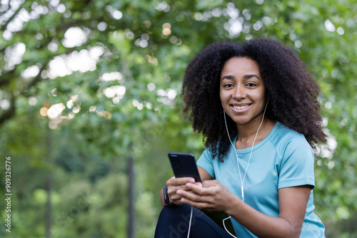 Young African American athlete smiling while using smartphone outdoors in park. Wearing earphones and blue sportswear, enjoying nature and technology.