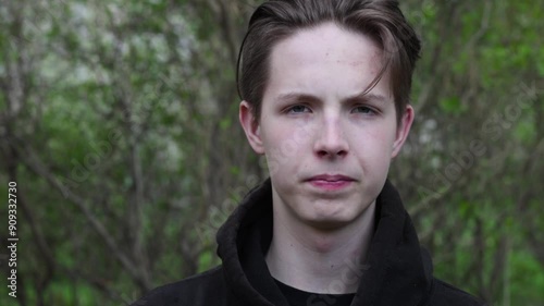 Close-up of a young boy's face against a background of branches. Cute teenage boy in a black hooded biker spitting sideways.High quality FullHD footage