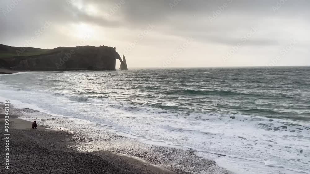 Vue sur les célèbres falaises d'Étretat, en Normandie, France. Le soleil se reflète sur l'eau, baignant la scène d'un halo de lumière magnifique. Quelqu'un observe les vagues accroupi depuis la plage.