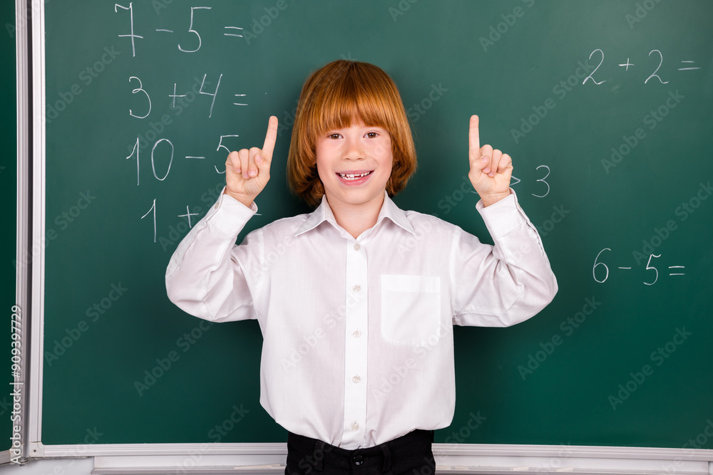 Photo of charming little pupil boy point up excited empty space wear trendy uniform classroom blackboard background Photo of charming little pupil boy point up excited empty space wear trendy uniform classroom blackboard background