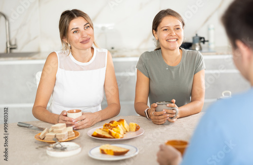 Smiling hospitable woman and young girl receiving male guest over cup of tea with sweets at home, sitting at cozy kitchen table and talking friendly..