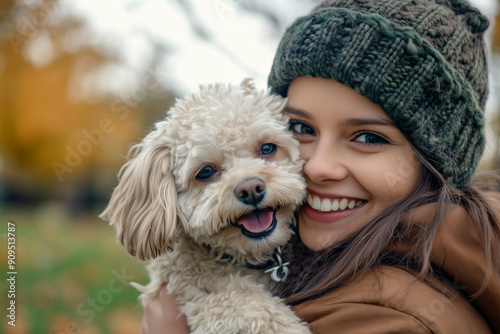 woman is holding a small white dog and smiling. The dog is wearing a collar and he is happy. The woman is wearing a green hat and a brown coat