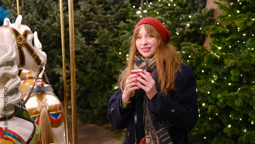 Beautiful young woman with red hair at the Christmas market near carousel in a red beret and coat drinks ,smiling and enjoying hot chocolate from a red cup.Hight quality photo