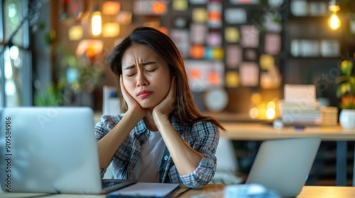 Young asian woman experiencing a headache while working in the office. Showing discomfort as she holds her head. strain from prolonged sitting and laptop use.